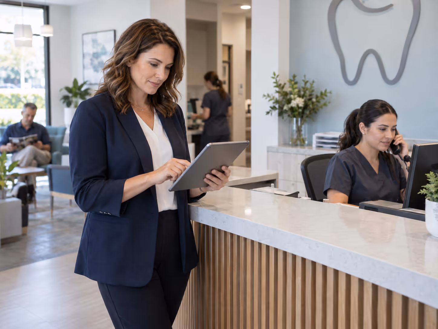 A dental practice manager reviews information on a tablet while the front desk handles a call.
